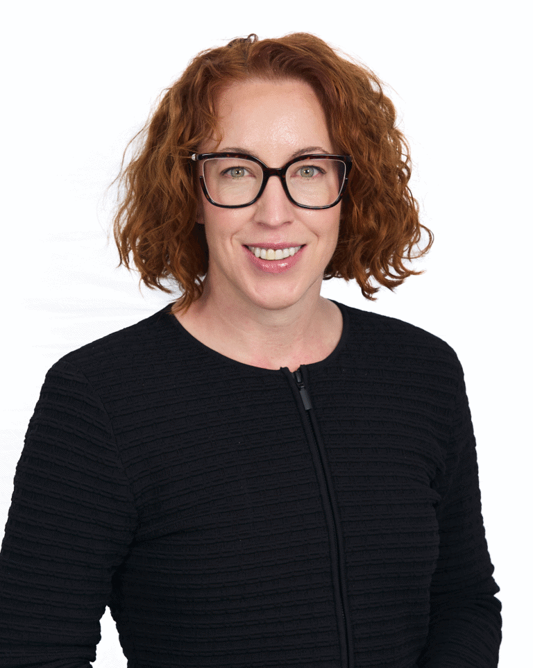 Professional woman with curly red hair and black glasses smiling against a white background, representing diversity and confidence in the workplace.