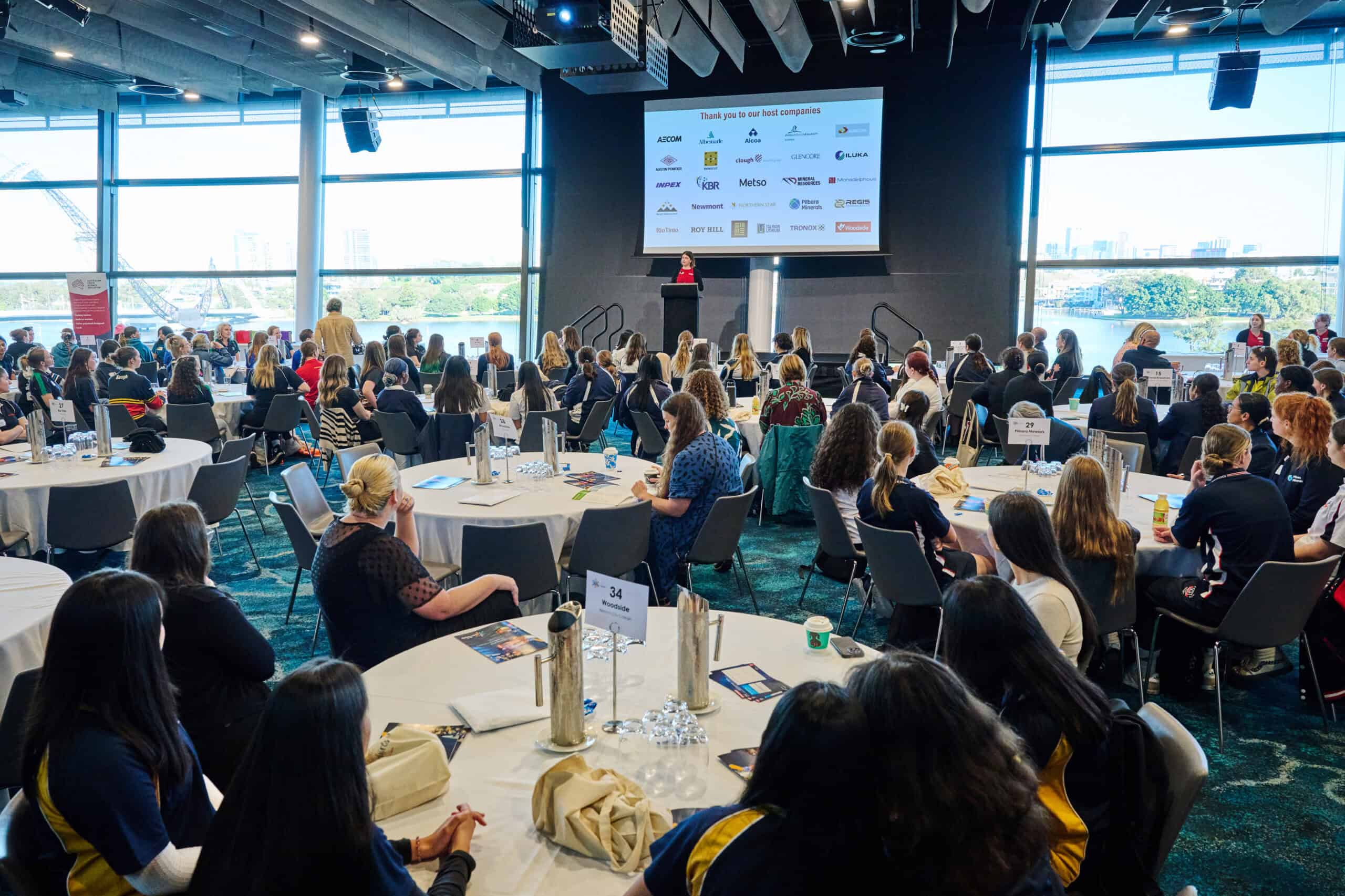CMEWA conference with diverse attendees listening to a speaker on stage, large windows showing city skyline and river, emphasizing professional networking and educational event in a modern venue.