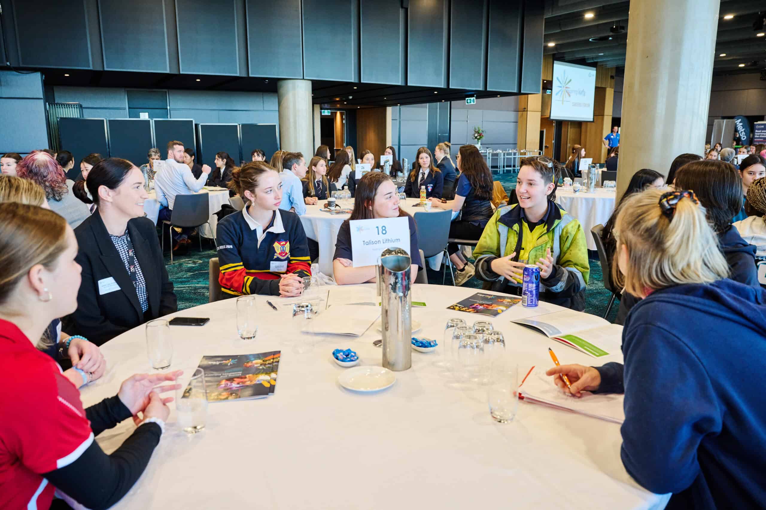 Students participating in a youth conference or educational event, sitting at round tables engaged in discussions, with a professional and collaborative environment, emphasizing youth engagement and leadership.
